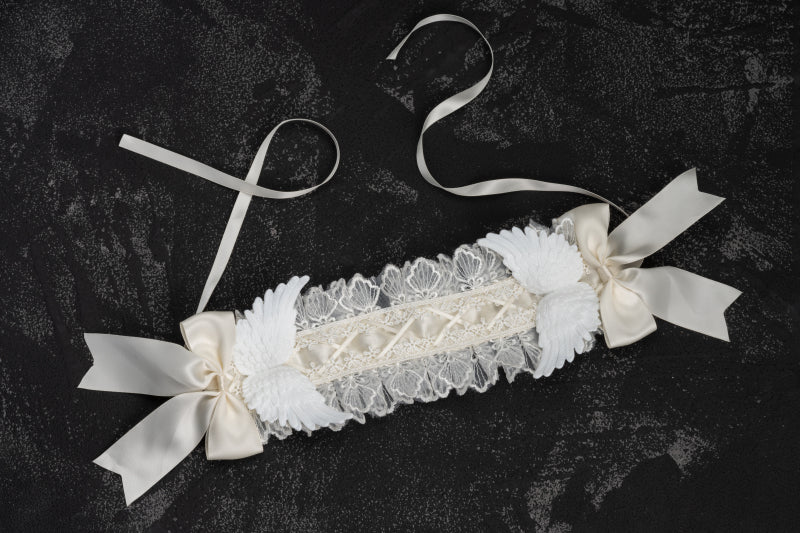 Flat lay overhead view of unworn gothic lolita white lace angel wings hairband on dark textured background, showing satin bows and adjustable tie ribbons