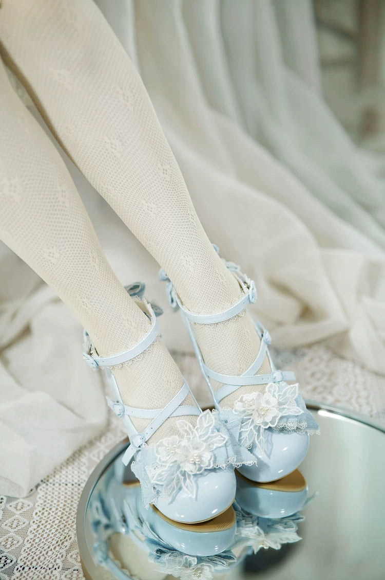 Light blue polished patent leather sweet Lolita heels in high-angle mirror reflection view showing model's feet in sheer fishnet stockings, 3D floral toe cap appliques and reflected sole