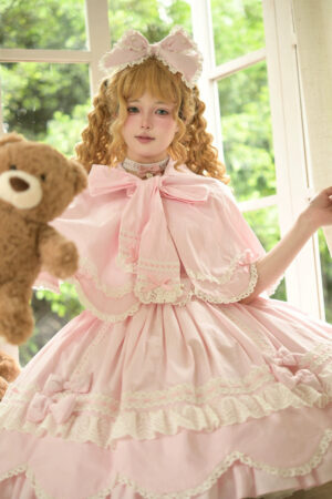 Pink vintage petal rabbit bunny ear lolita cape front three-quarter view on curly blonde lolita model holding teddy bear in sunny bedroom with white lace trims and large front bow