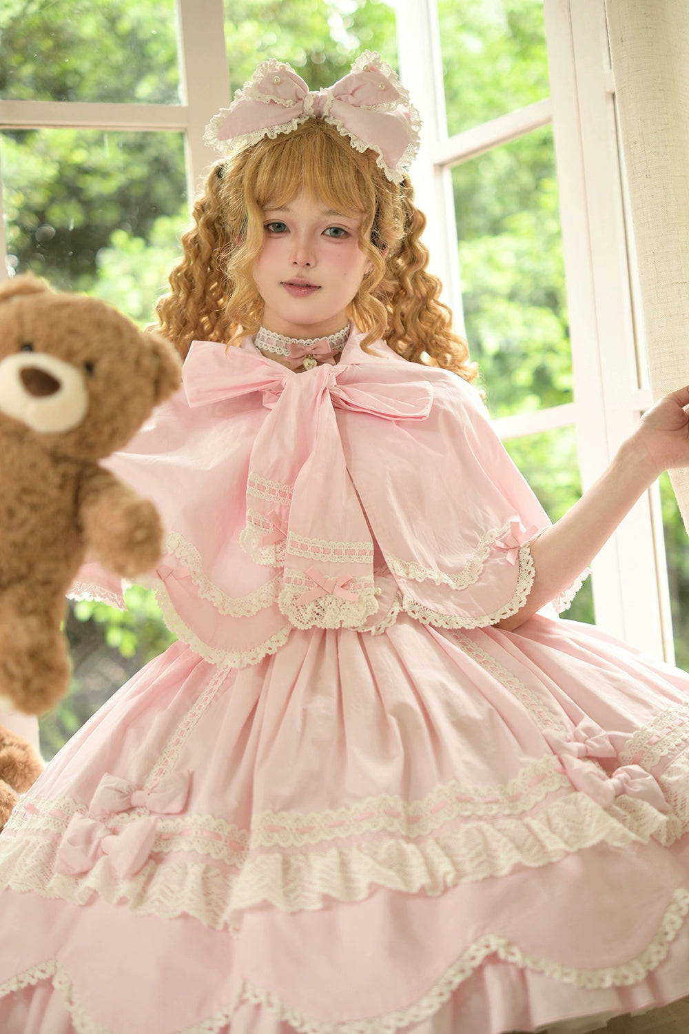 Pink vintage petal rabbit bunny ear lolita cape front three-quarter view on curly blonde lolita model holding teddy bear in sunny bedroom with white lace trims and large front bow