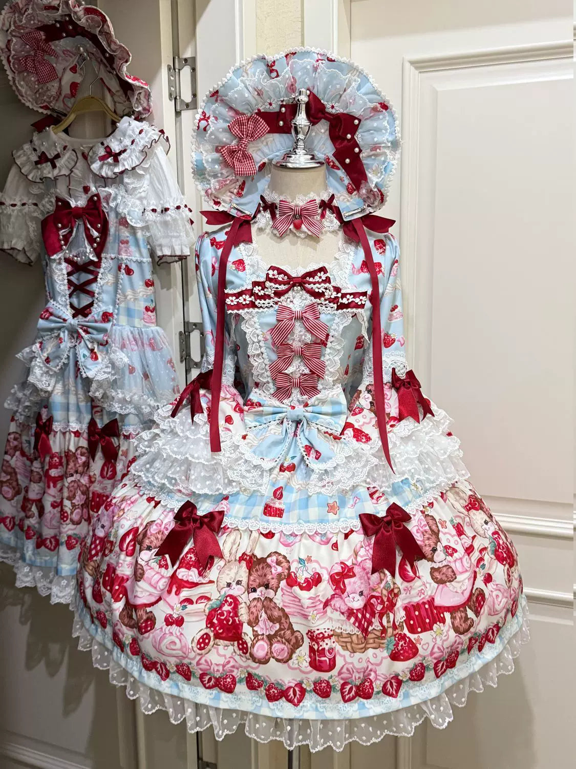 Two lolita OP dresses on mannequins featuring blue gingham and red strawberry bear print bases, both with square necklines, lace trims, big bows, and matching bonnets