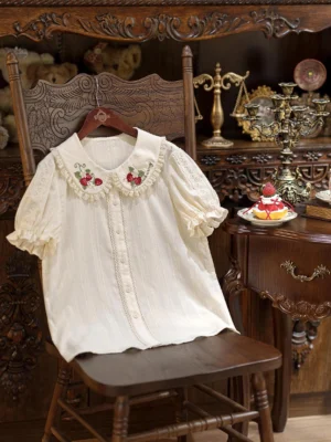Flat lay hanging view of cream lolita cotton blouse with red floral strawberry embroidery on ruffled Peter Pan collar and puff short sleeves