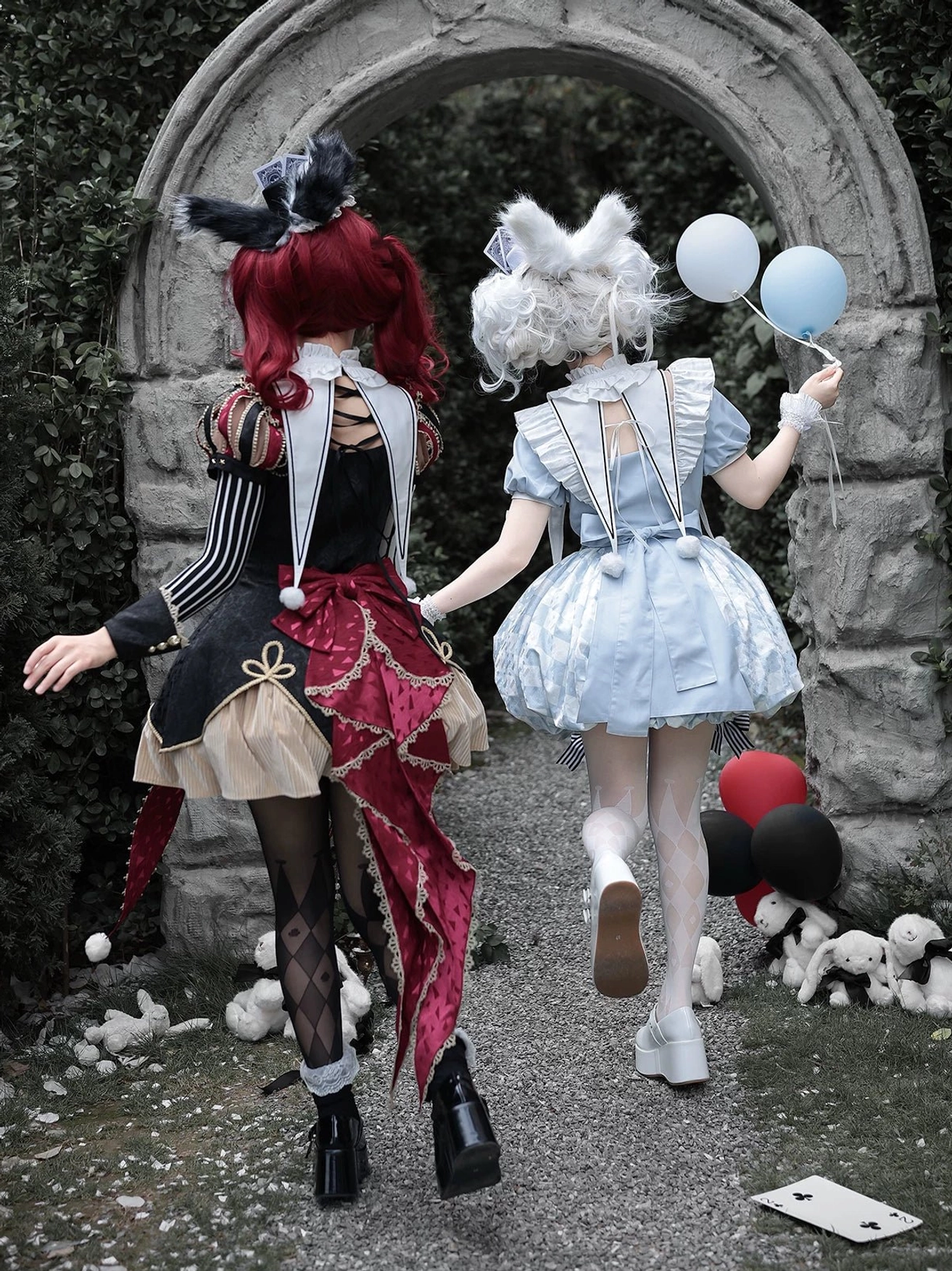 Back view of blue Alice Lolita OP dress showing open back design and bubble hem construction, paired with red-black themed dress rear detail in stone garden with balloon props