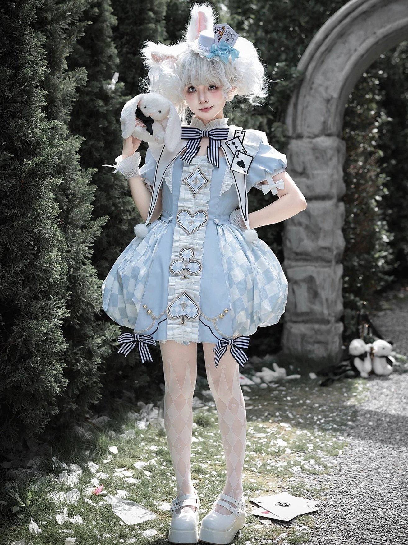 Dreamy blue Alice-inspired Lolita OP dress with bubble hem, heart and playing card embroidery, and detachable striped bow, worn with rabbit ear hair accessory in vintage stone garden setting