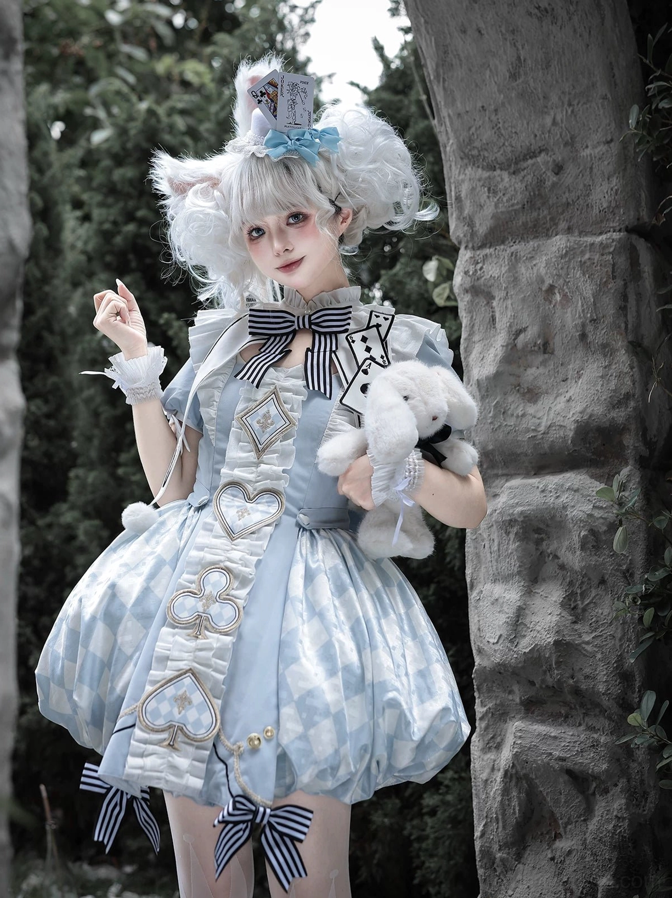 Close-up of model wearing blue Alice Lolita OP dress holding plush rabbit toy, detail view of ruffled wrist cuffs, chest playing card badge, and classic bubble hem silhouette