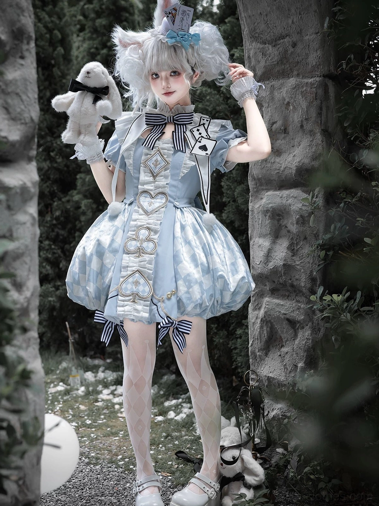 Three-quarter front view model adjusting hair ornament wearing blue Alice Lolita OP dress, clear showcase of dress silhouette, chest embroidery, bow tie details in outdoor stone setting