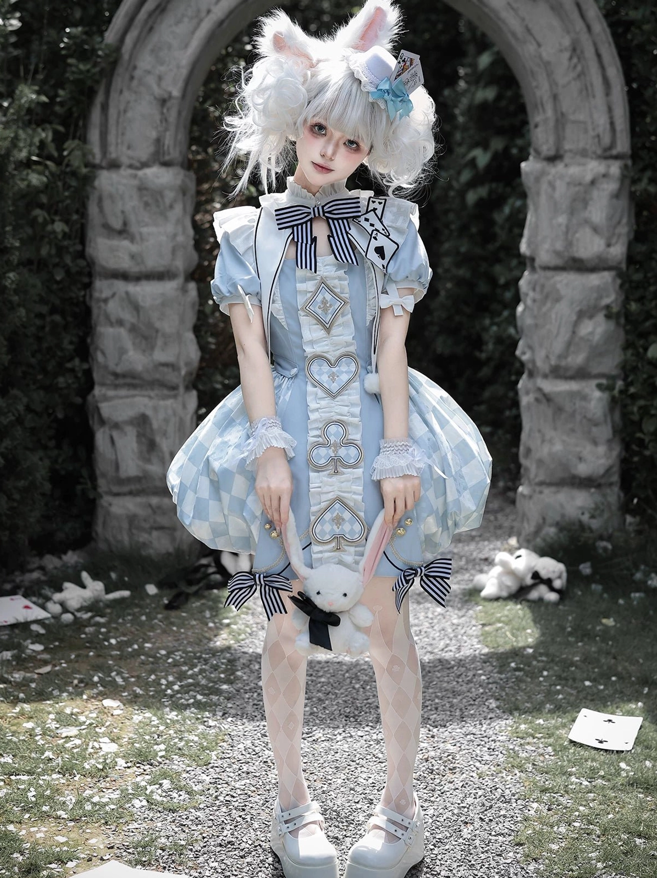 Front full-body view of model wearing blue Alice Lolita OP dress with plush rabbit toy, classic bubble hem construction, striped detachable bow at hem, stone garden background with props
