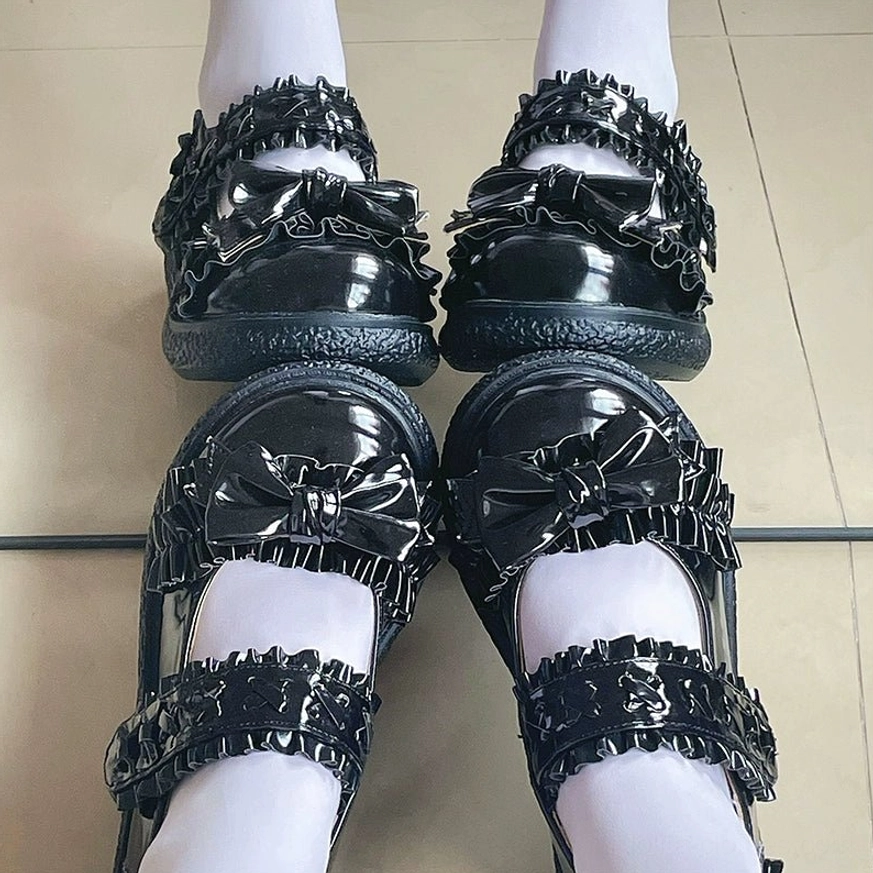 Overhead flat lay display of all-black Lolita Mary Jane flat shoes on tiled floor, full black patent leather with ruffled straps and bow-shaped toe decor