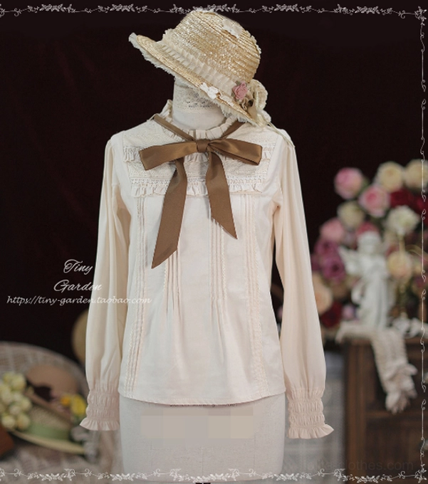 Front full view of apricot beige Lolita long-sleeve blouse on dress mannequin with flower-decorated straw hat, showcasing brown fabric bow necktie, ruffled stand collar, and front pin-tuck pleats against retro floral studio background