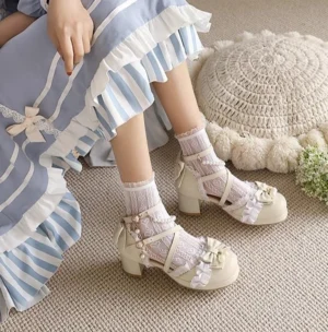 Side close-up detail of apricot Lolita Mary Jane sandals with round toe, ankle strap pearl chain, and bow embellishments worn on linen carpet floor with decorative crochet elements
