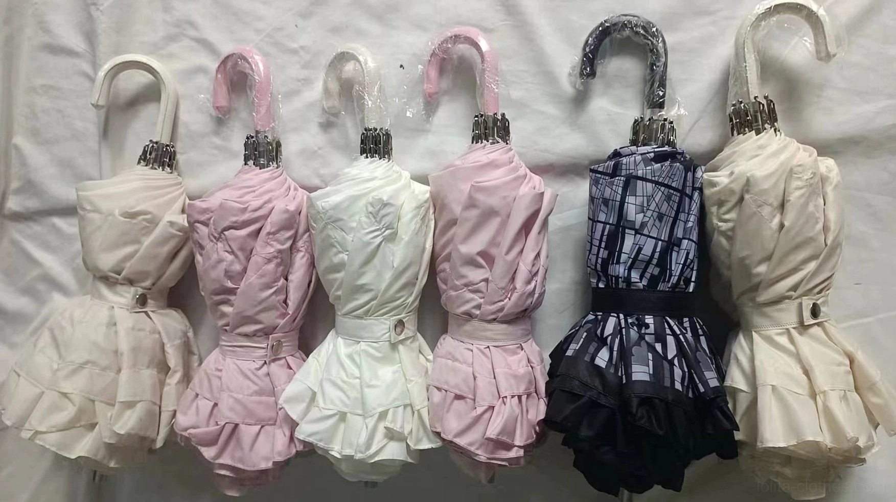 Assorted foldable Lolita parasols in multiple styles and colors including beige, pink, and black plaid with decorative dress-like covers on white cloth, overhead flat lay display