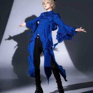 Male model wearing royal blue asymmetrical Ouji Lolita long coat with irregular hemline and flared ruffled princess sleeves, standing in dramatic studio spotlight with theatrical shadow backdrop