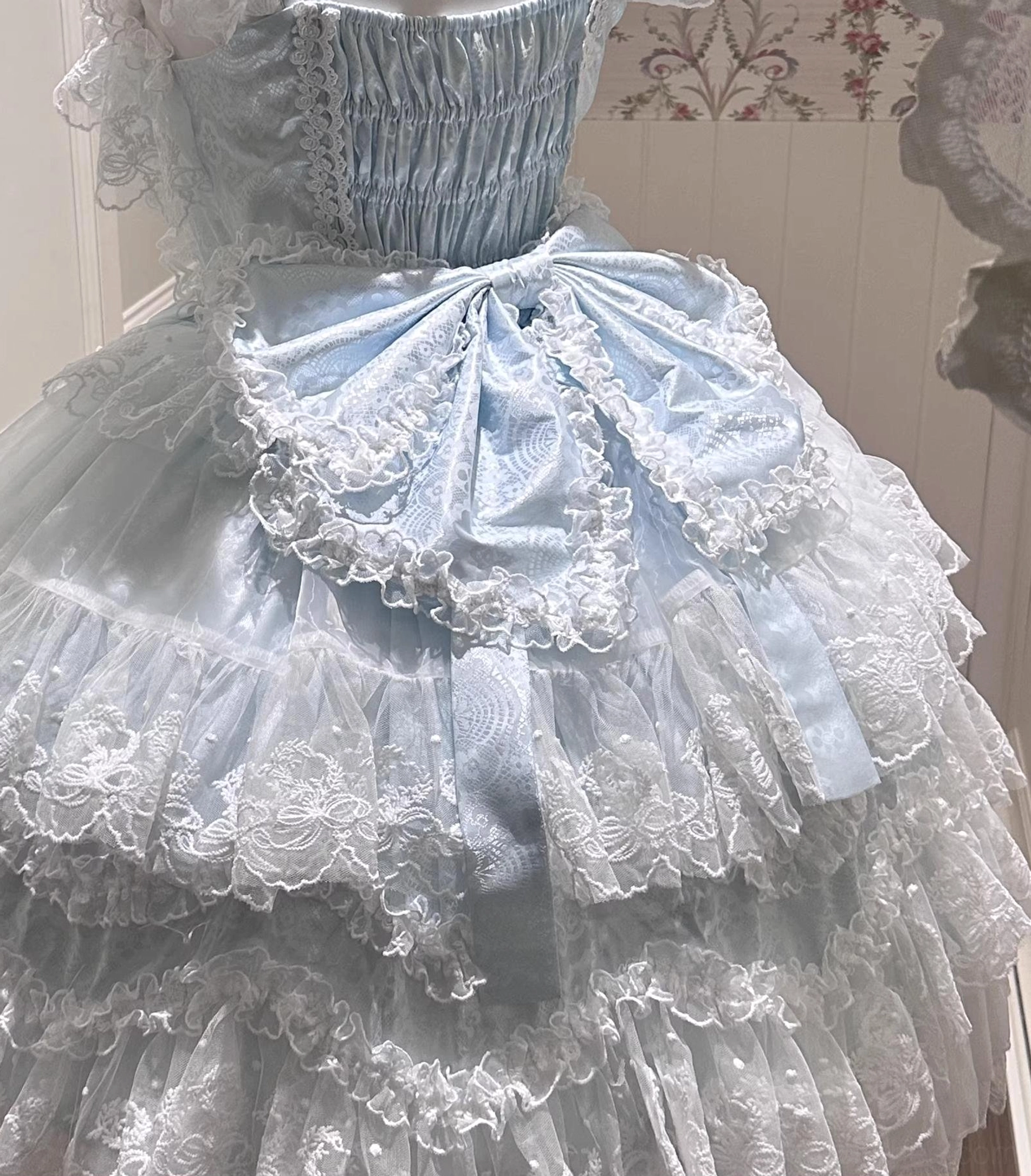 Back view close-up of baby blue Sakura Blossom Dream Sweet Lolita dress showing large layered lace back bow with pearl embellishments, shirred elastic waist construction, multiple layers of frilly tulle skirt with delicate lace trim, demonstrating traditional Sweet Lolita architectural silhouette