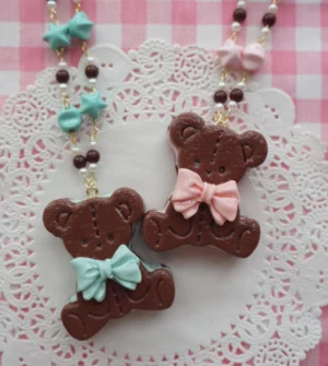 Bear cookie clay pendant necklace flat lay featuring dark brown bear with bow tie on pink gingham tablecloth with white lace doily, colorful star beaded chain with gold links