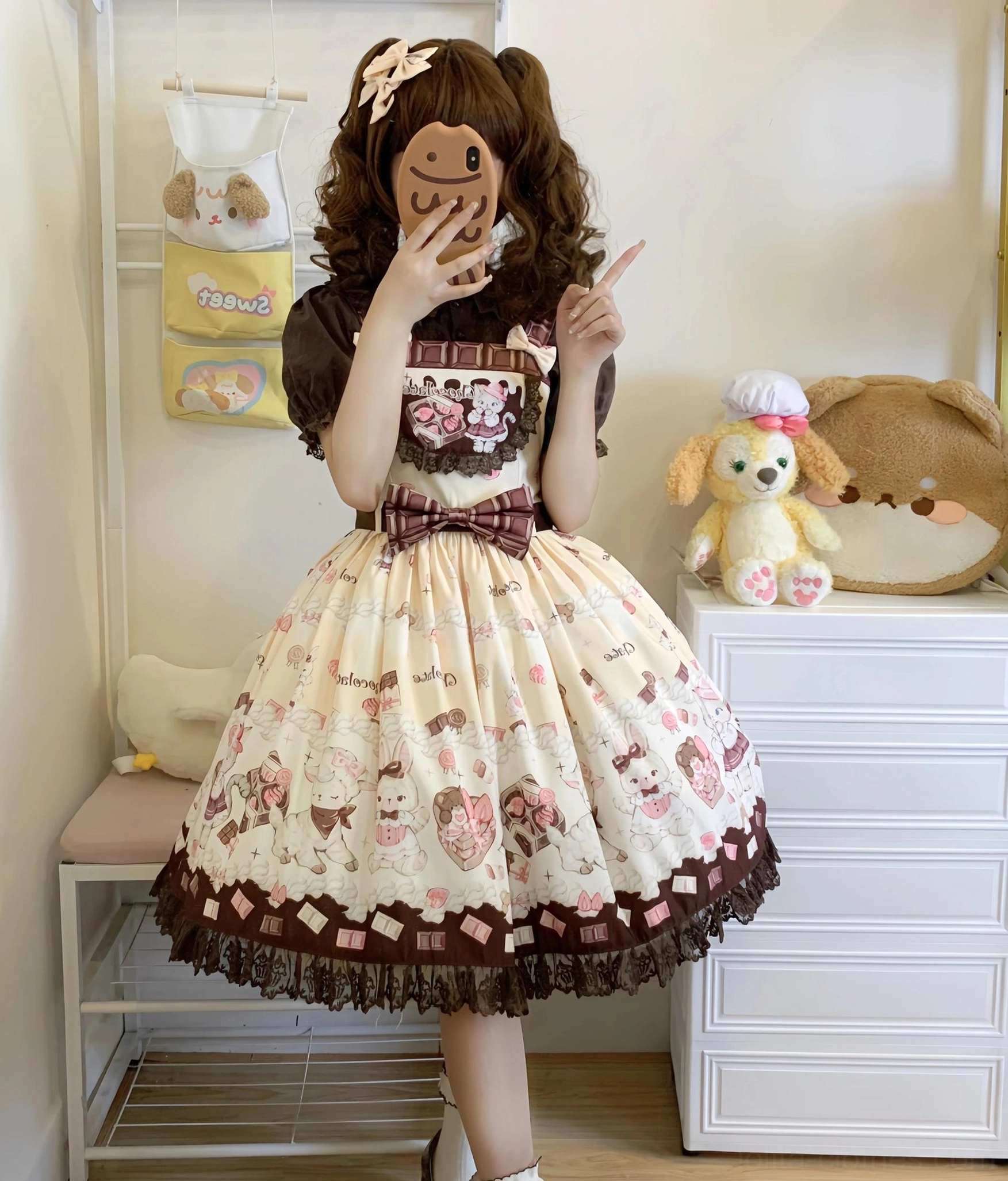Brown curly-haired twin-tailed model taking hidden face selfie wearing classic beige and brown cat print Lolita salopette dress with front waist bow, detailed chest print, and lace skirt trim