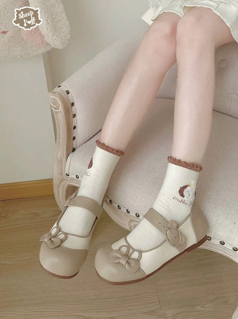 Close-up side view of beige bunny ear lolita flat shoes worn with embroidered frilled socks and linen armchair showing bear buckle strap and smooth leather upper