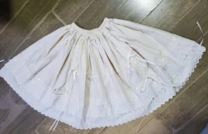 Beige cotton Lolita petticoat flat lay from above on wooden floor, showing elastic drawstring waist, full gathered circle skirt silhouette, and scalloped eyelet lace hem with decorative satin ribbon bows