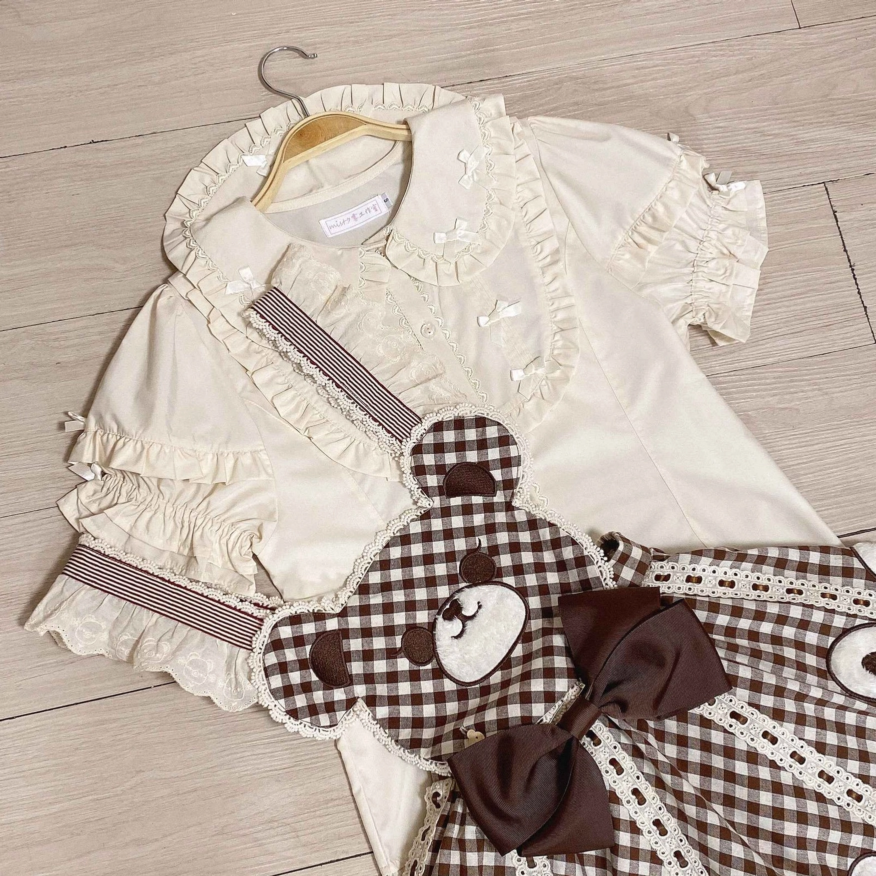 Beige lolita blouse with detachable gingham teddy bear apron striped decorative straps and layered ruffled sleeves flat lay wooden floor