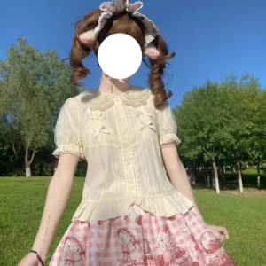 Beige sweet Lolita blouse with Peter Pan collar and puffed sleeves worn by model with twin-tail hairstyle in outdoor green lawn setting with matching pink gingham skirt