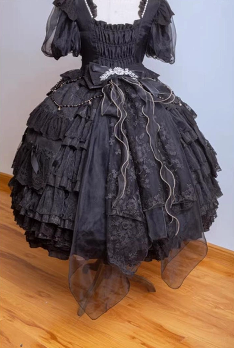 Black classic lolita OP dress flat lay close-up showing tiered lace skirt, short puff sleeves, square neckline, waist bow with rhinestone decor, and sheer tulle trailing hem with silver chain accents
