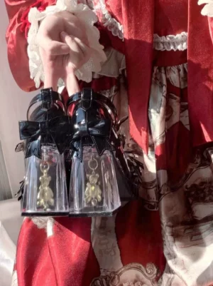 Black sweet lolita high heels with clear block heel containing tiny embedded bear, held by hands wearing red frilled lolita dress, overhead close-up shot, gold hanging bear charm, ankle bow decoration, transparent heel design