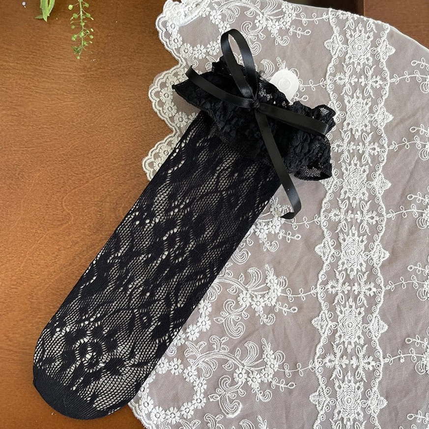 Single black French Lolita mid-calf lace sock overhead display showing sheer floral lace construction, ruffled cuff with black satin bow on embroidered lace placemat