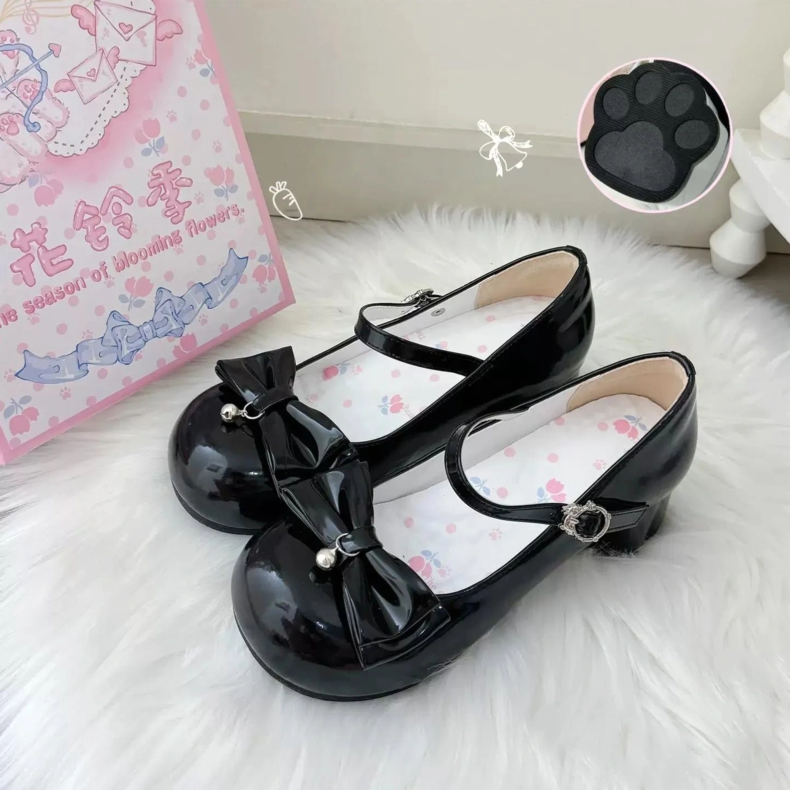 Black glossy patent leather sweet lolita Mary Jane shoes overhead close-up view showing large front bow decoration, silver ankle buckle strap, chunky block heel structure, cat paw-shaped heel inlay detail sample, white fur rug display with branded packaging