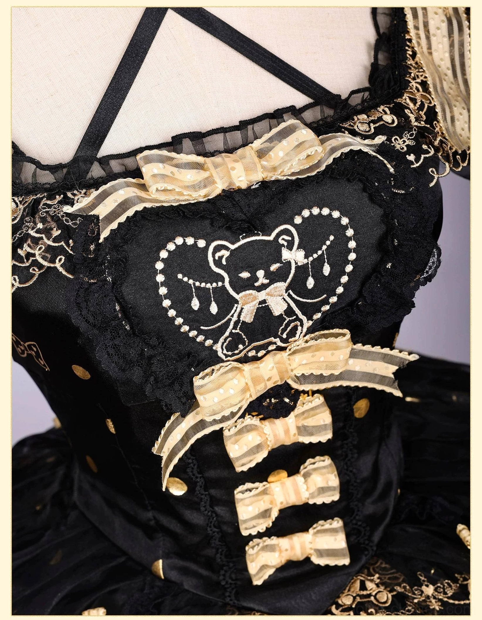Close-up detail of lolita dress front bodice featuring heart-shaped bear embroidery, stacked gold satin bows, delicate lace trims, cross shoulder straps on black fabric with tiny gold dot prints