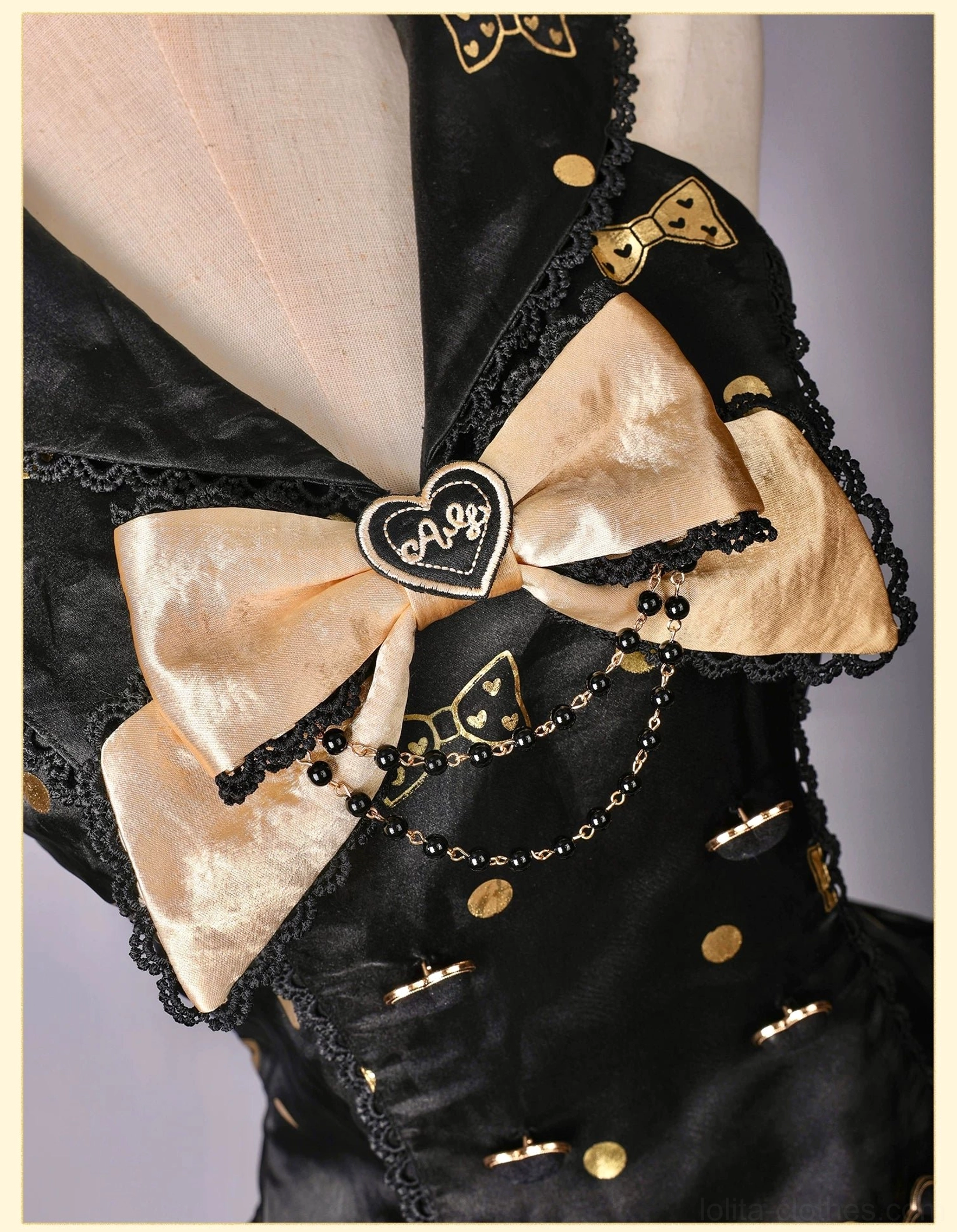 Detailed close-up of lolita dress upper bodice showing large gold satin bow with heart-shaped metal charm, bead chain decoration, tiny gold motifs, and delicate lace neckline trim