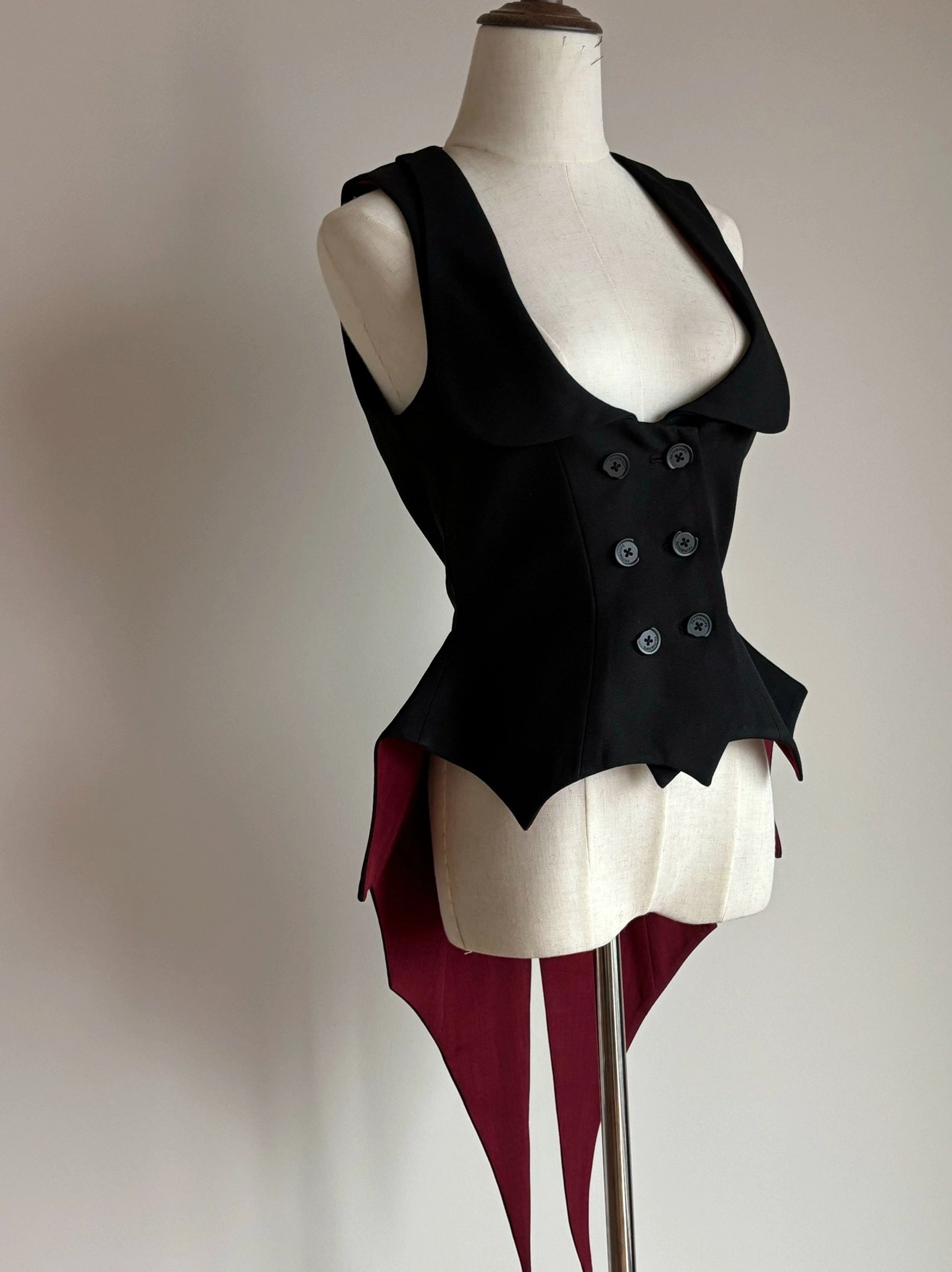 Black Gothic Lolita vest with wine-red bat-wing hem displayed on dress form, left three-quarter front view showing double-breasted button closure and deep U-neckline
