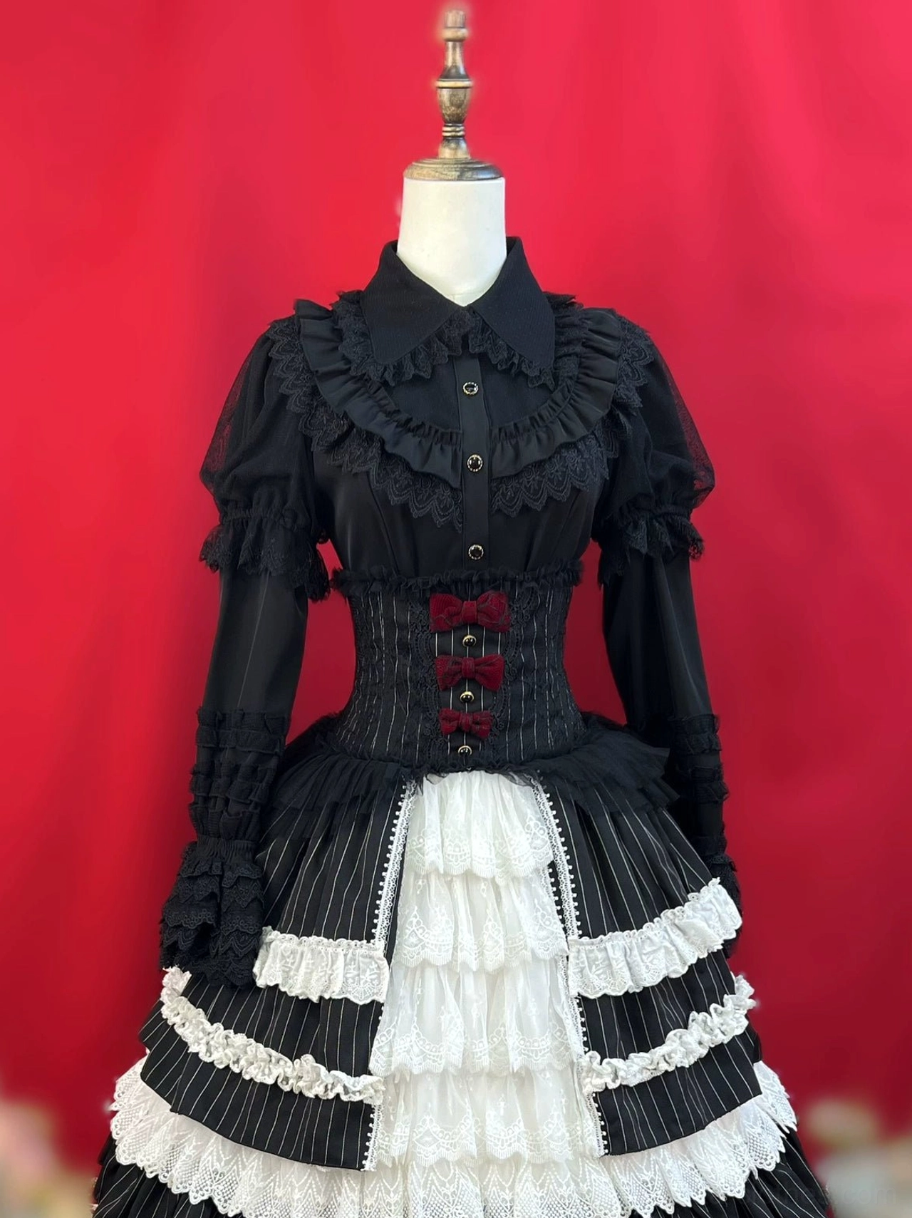 Black gothic lolita blouse with matching corset skirt set featuring puffed ruffled long sleeves front button closure striped corset waist bow accents and layered ruffled lace hem on dress form red studio background