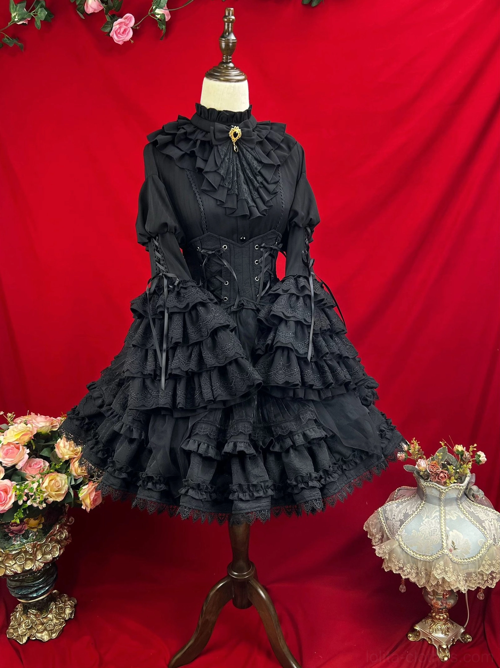 Black gothic lolita dress displayed on mannequin showing tiered ruffled sleeves high neck ruffled collar corset waist and layered ruffled skirt against red velvet backdrop