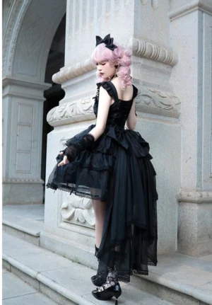 Black Gothic Classic Lolita high-low JSK dress side back view showing dramatic bow trailing and tiered ruffled skirt, model wearing matching lace accessories in vintage European stone arch outdoor location