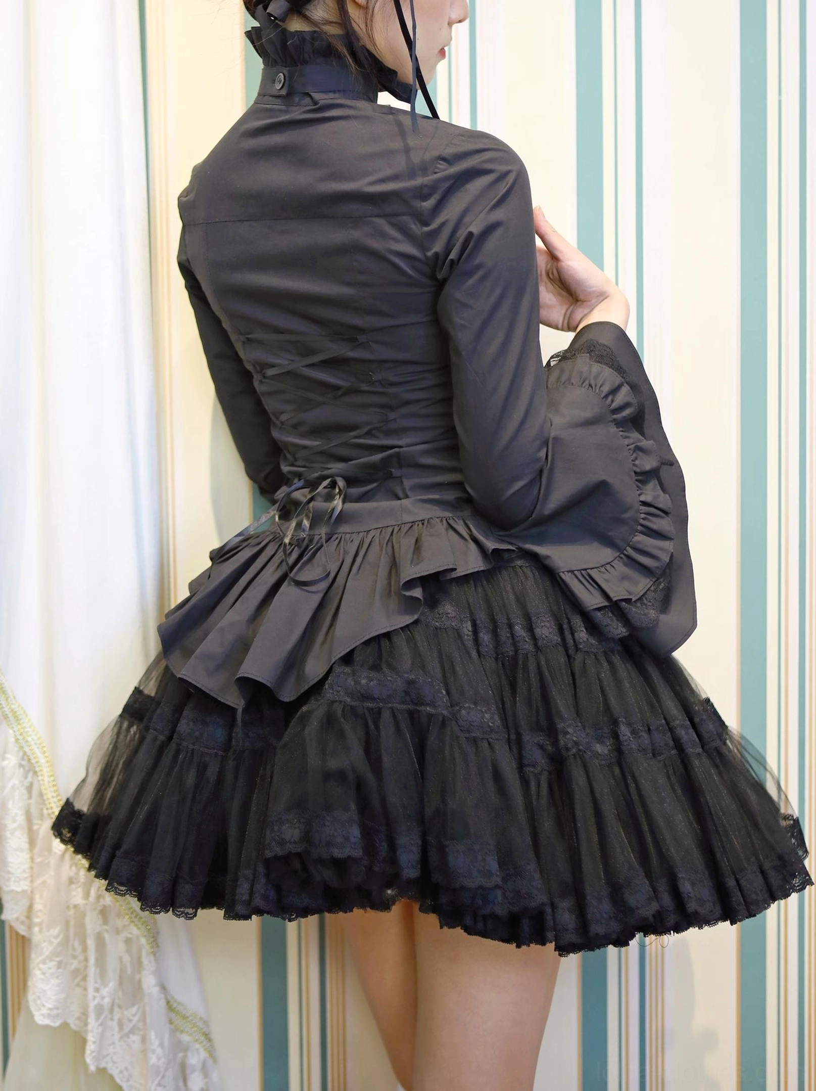 Black gothic lolita ruffled blouse back three-quarter view, lace-up back closure ruffled tiered hem flared trumpet sleeves fitted silhouette on model