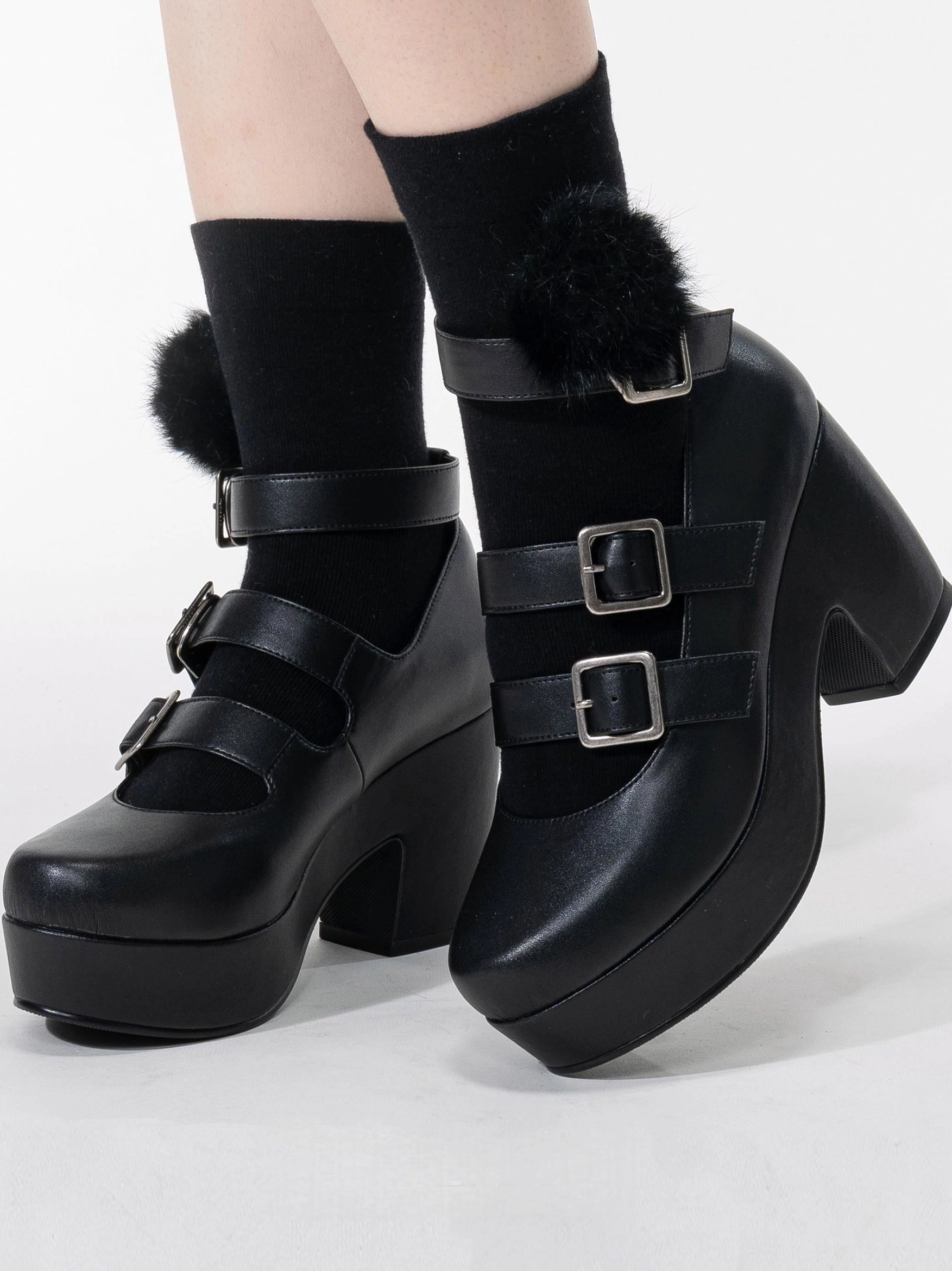 Black triple-buckle Gothic Lolita platform heel shoes with matte smooth leather, three adjustable silver buckle straps, chunky block heel, and fluffy pom-pom decorated black socks on model feet