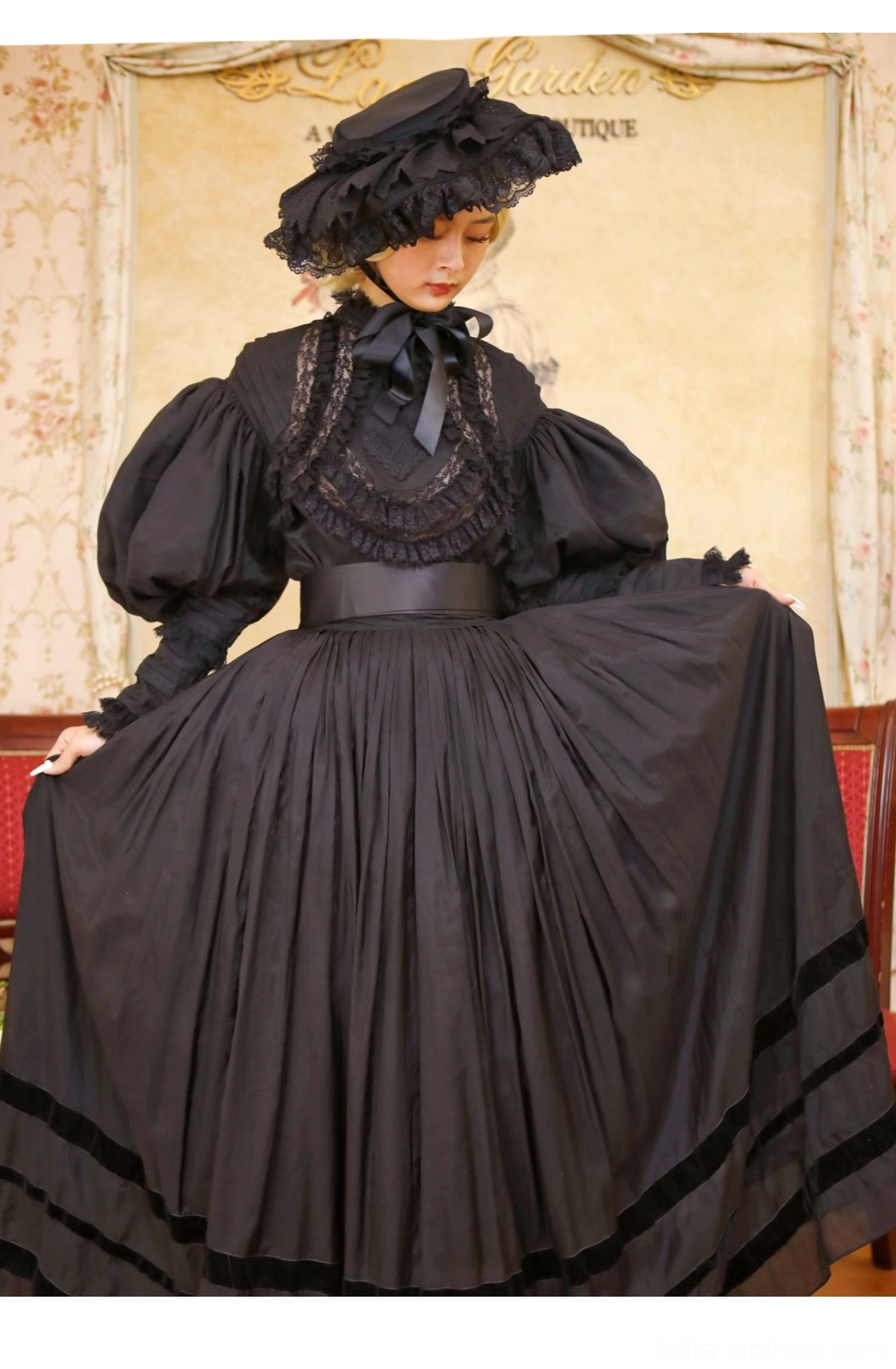 Black Gothic Victorian Lolita dress front three-quarter view on seated model featuring ruffled high collar with lace overlay, giant puff sleeves, wide waist belt, pleated long skirt with velvet hem trim