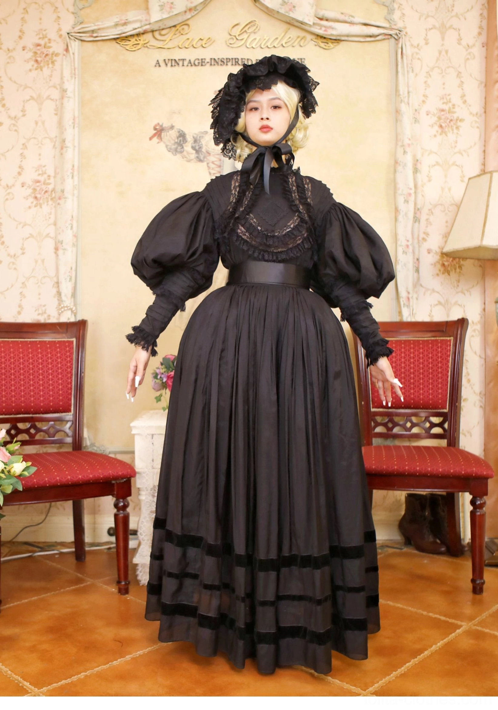 Full front standing view of model in black Gothic Victorian Lolita formal dress wearing vintage black bonnet, lace neckline with satin bow, oversized puff sleeves, wide waist belt, and long pleated skirt with velvet hem trim