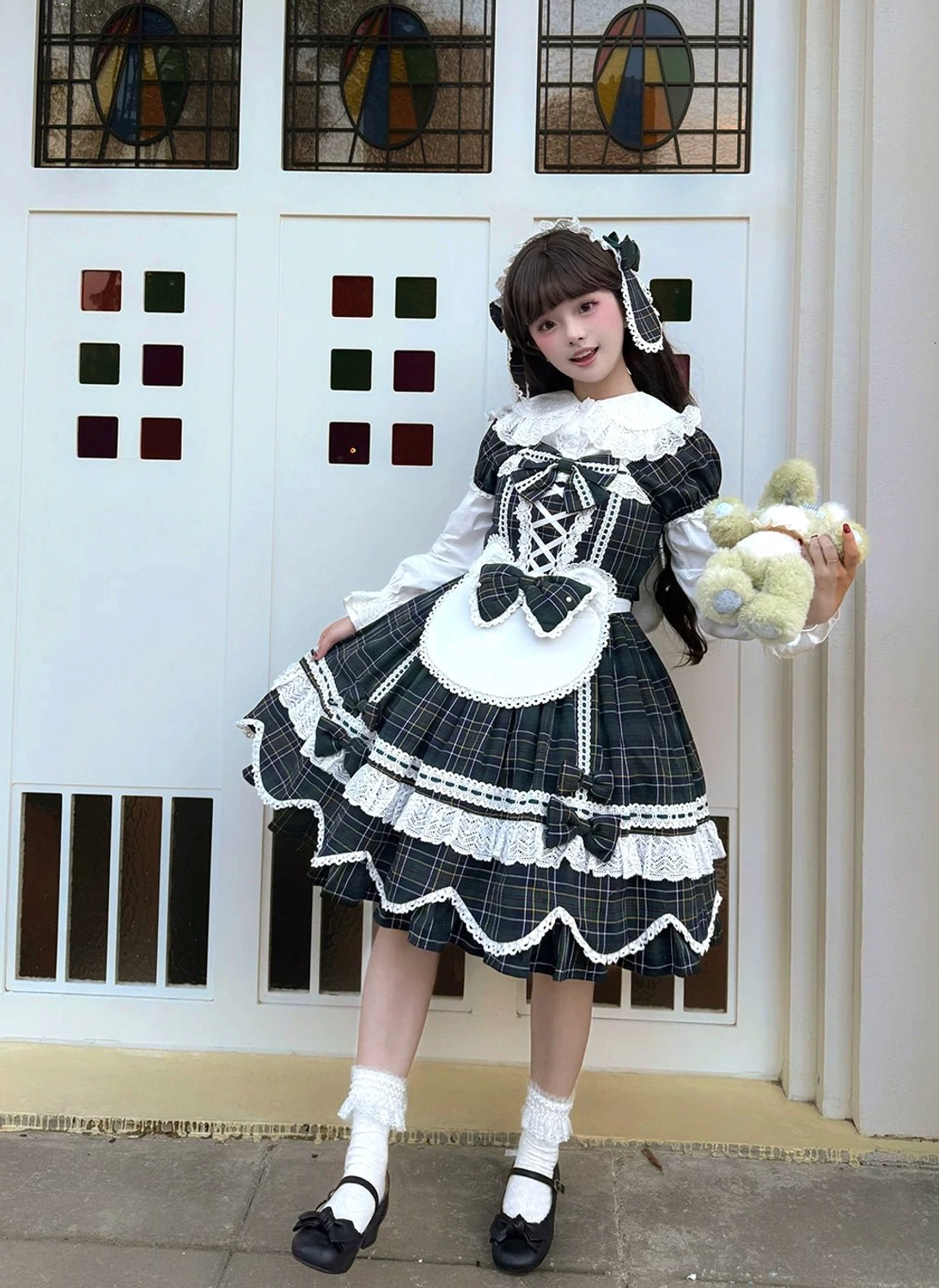 Black-gray tartan plaid maid-style lolita OP dress with white apron front, ruffled white under-sleeves, matching hair clips, door background indoor setting