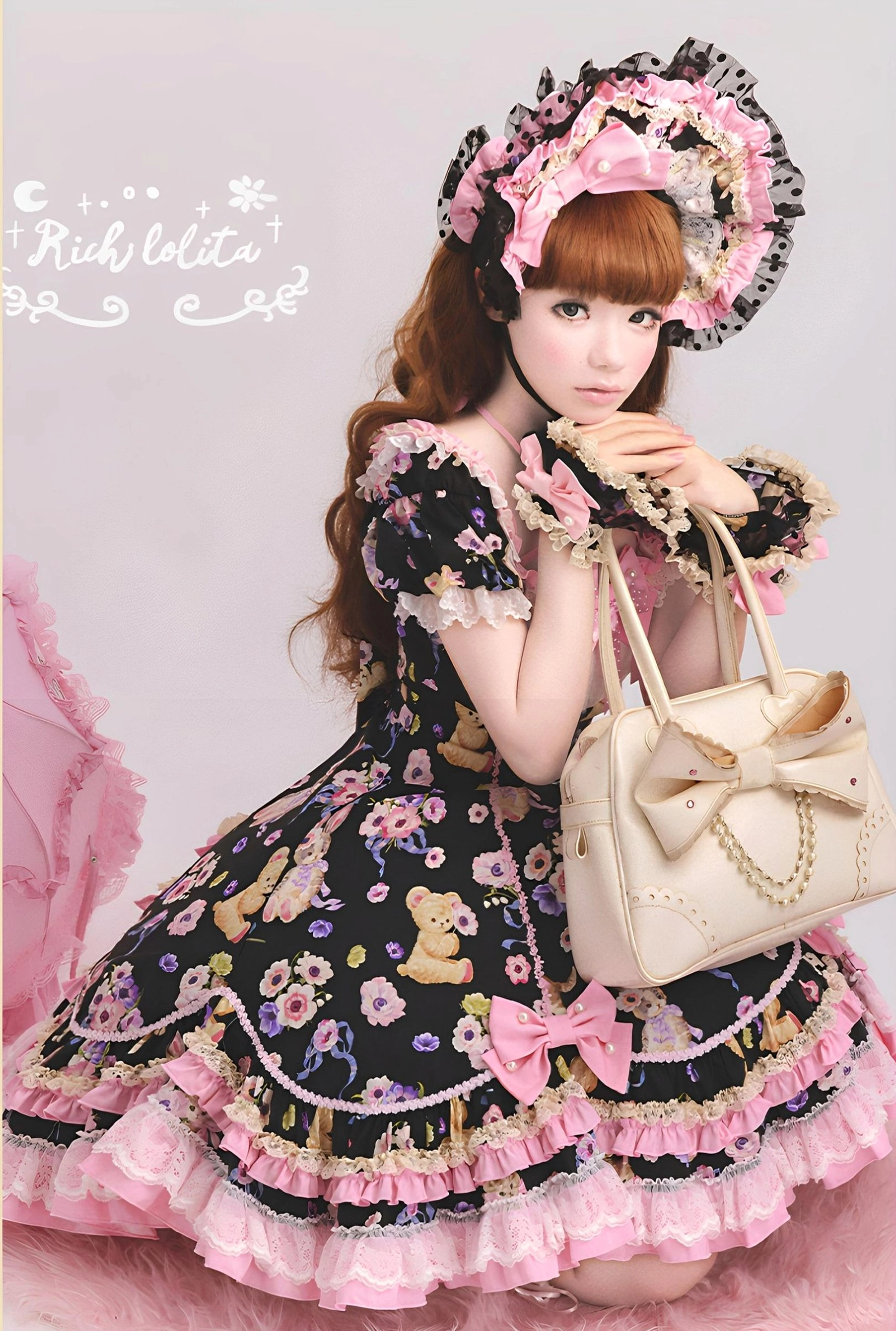 Full-body kneeling view of black sweet lolita OP dress with complete matching set, lace bonnet, pink ruffled hem, beige bow-handled handbag, teddy bear floral prints, pink studio rug