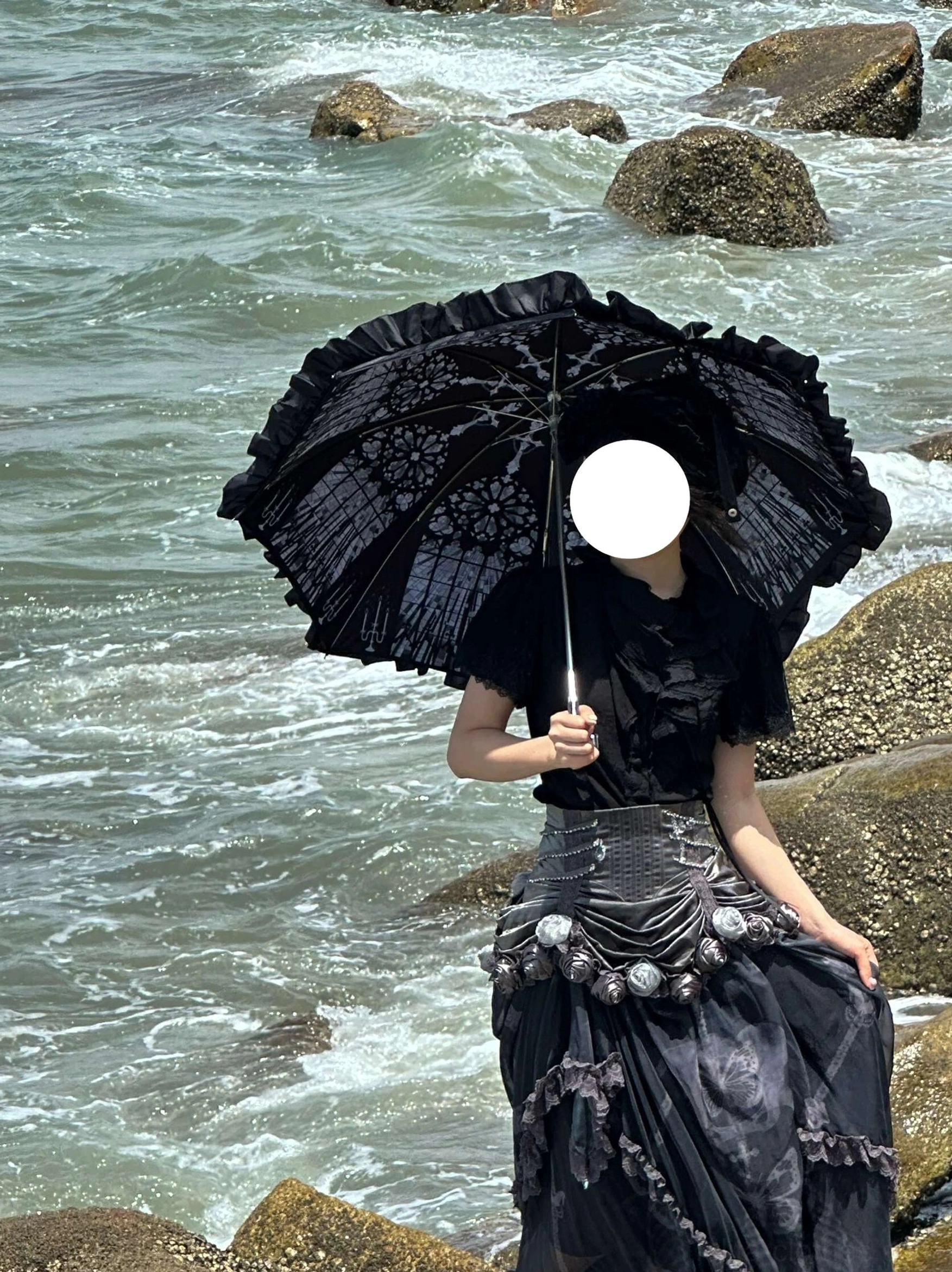 Model in black gothic Lolita gown holding black lace-print parasol with ruffled canopy at rocky seaside, front view demonstrating gothic coordinate styling