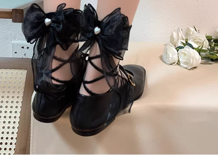 Front full lower leg and foot view of pair of black lolita ankle socks with dual pearl-centered organza bows and sheer straps worn with black leather Mary Jane shoes on white rose decorated table