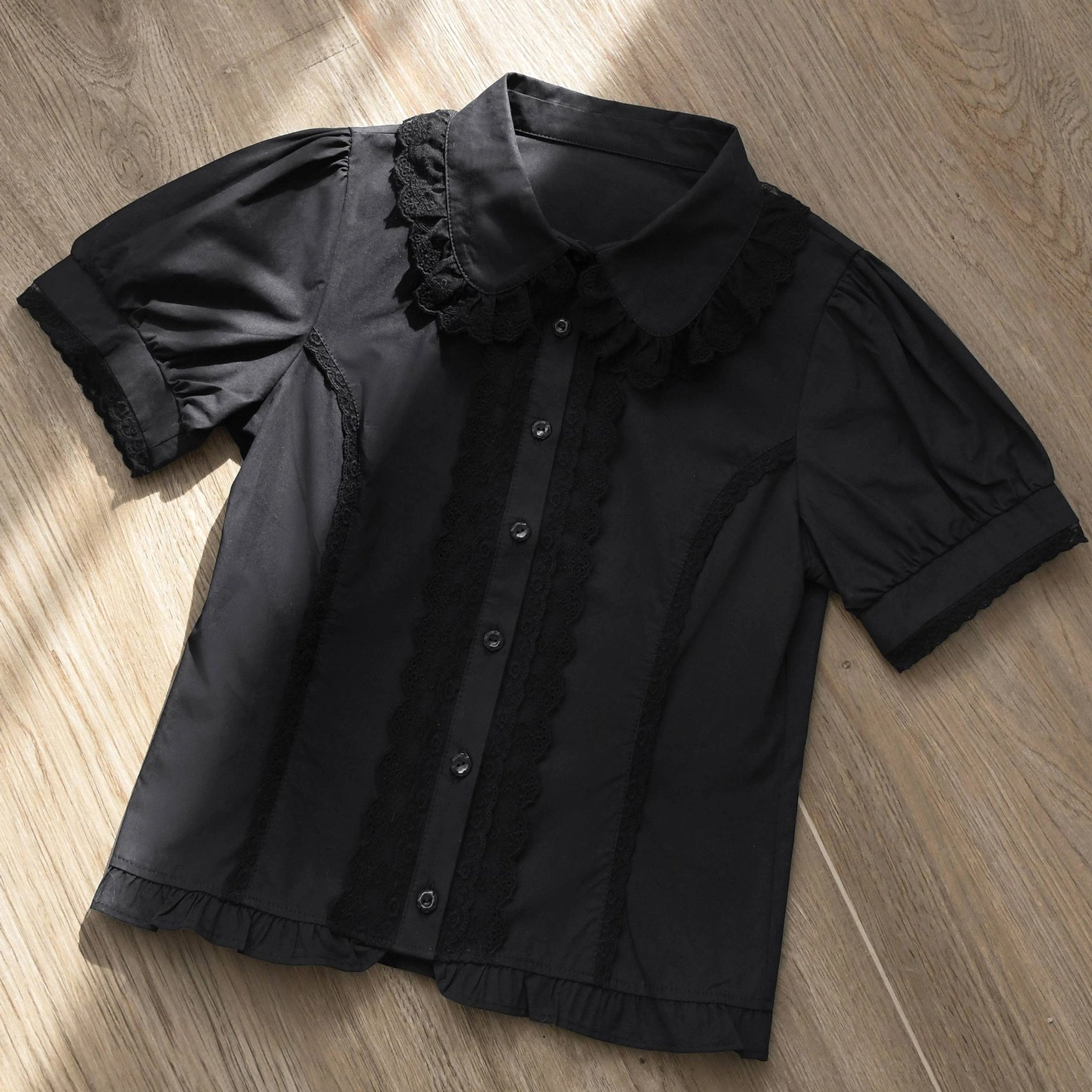 Black vintage Lolita short-sleeve blouse with black lace ruffled collar button front puff sleeves and ruffled shirt hem flat lay wooden background