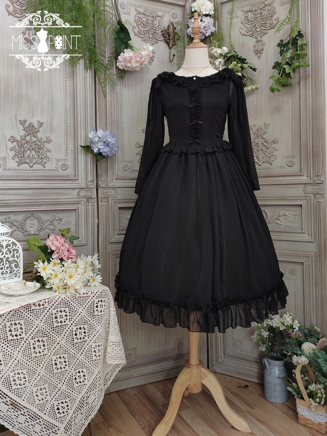 Pure black lolita one-piece dress with ruffled crew neckline, dimensional ruffled waist trim, and full pleated skirt with ruffled hem, displayed in sheer chiffon fabric on dress mannequin, vintage indoor room with lace tablecloth decor