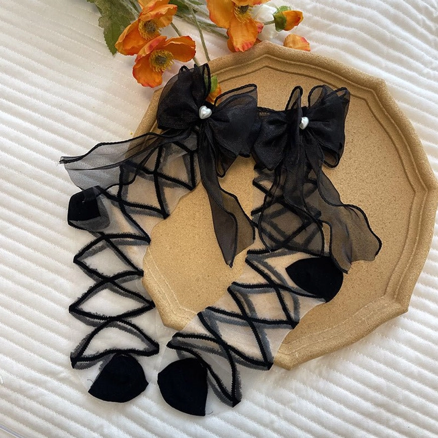 Overhead flat lay of black lolita gauze ankle socks with crisscross sheer straps, pearl-studded organza bows and velvet heart-shaped accents on decorative tray with floral props