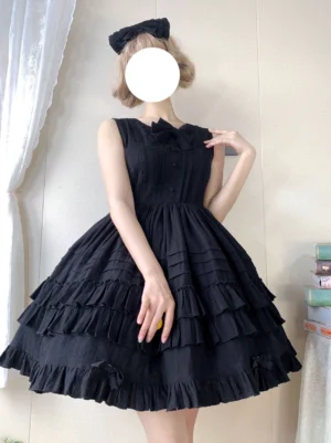 Black classic lolita JSK jumper dress with tiered ruffles and chest bow worn by female model in indoor setting showing front three-quarter view