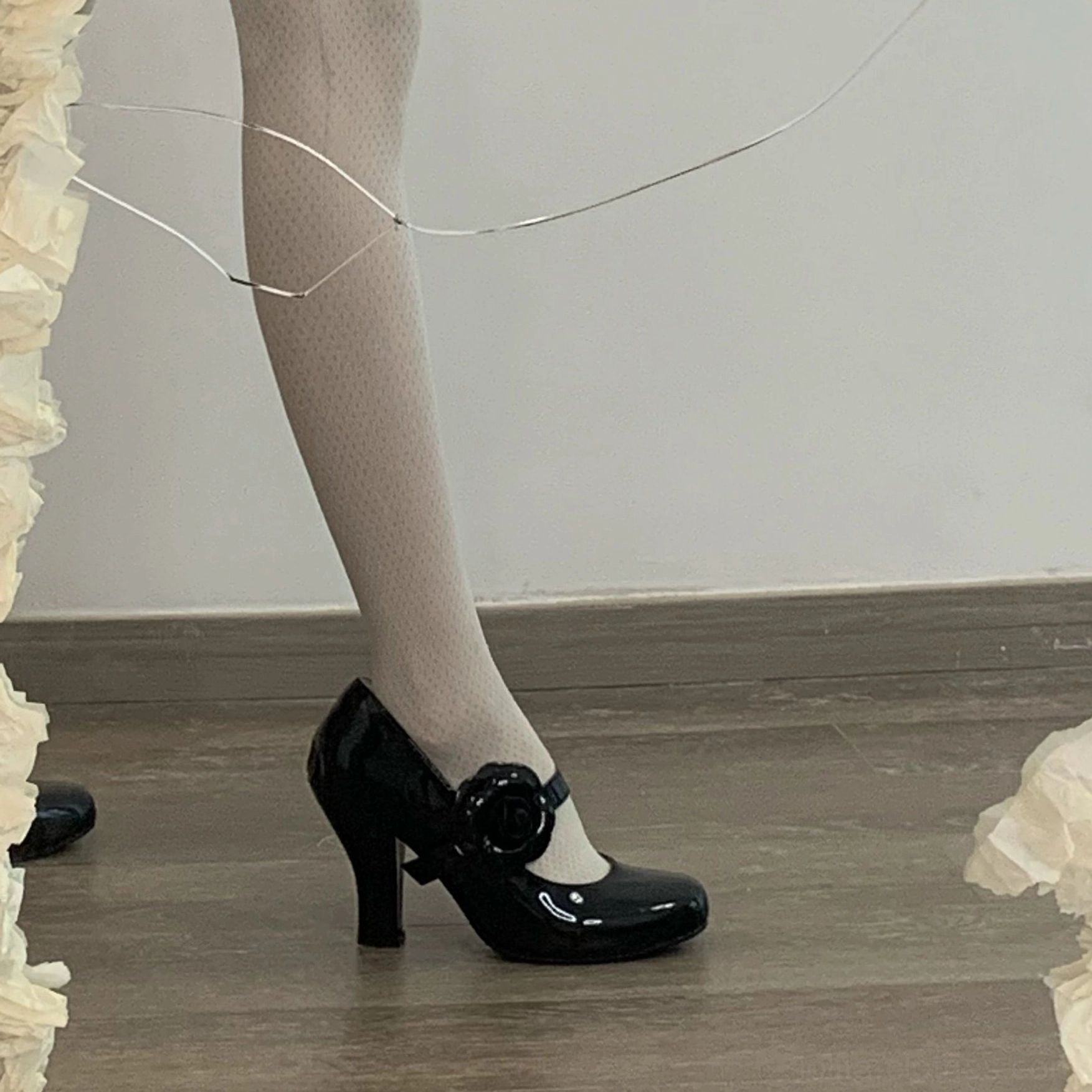 Black patent leather Lolita Mary Jane high heels side view of wearing foot wooden floor with fairy lights ruffled decor block heel rose ornament ankle strap sheer dotted tights