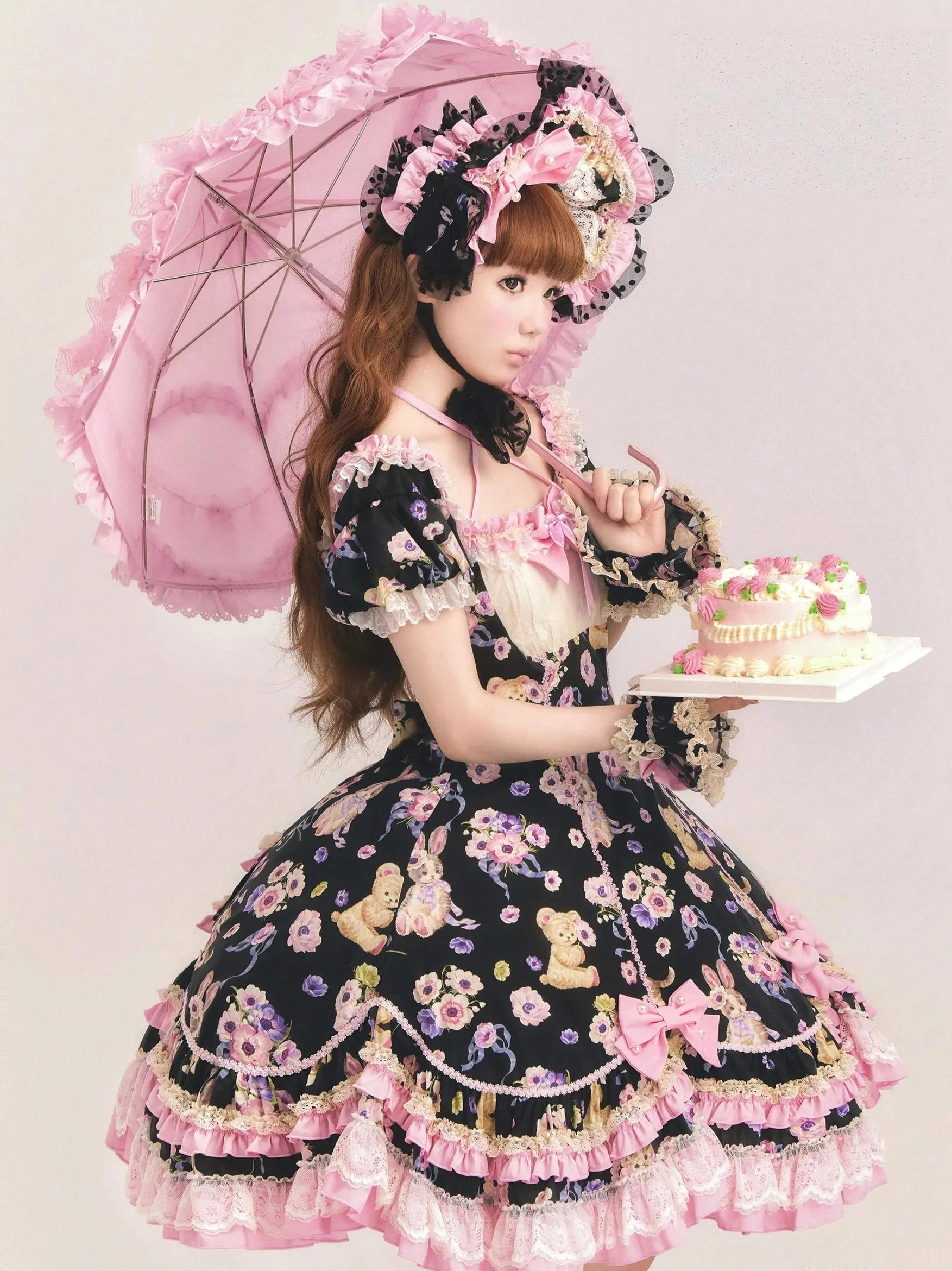 Three-quarter side view of black sweet lolita OP dress with matching bonnet and pink ruffled hem, teddy bear floral prints, model holding pink lace parasol and cake tray, studio photography