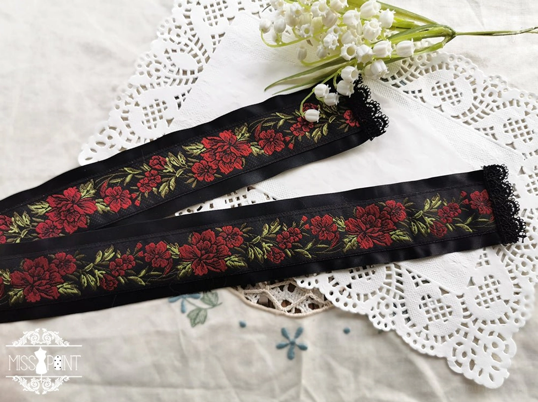 Overhead flat lay view of black satin lolita ribbon belt with red peony embroidery and pearl-beaded lace trims on ornamental lace background