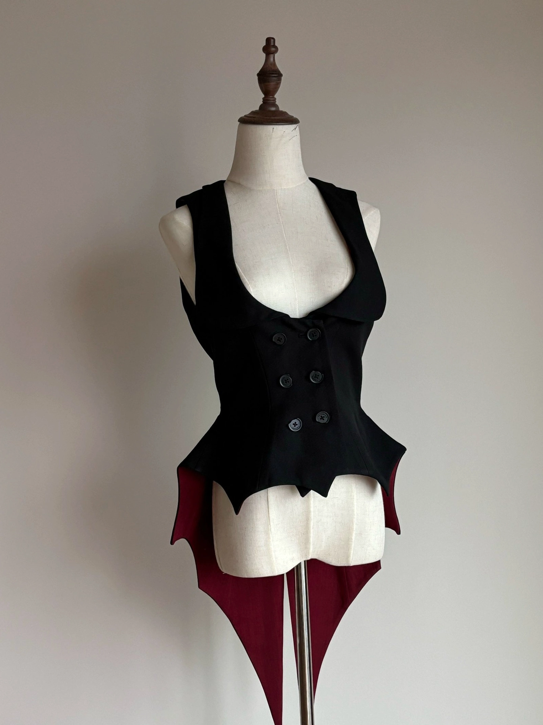 Black sleeveless Gothic Lolita waistcoat with asymmetrical wine-red bat-wing tail hem shown directly from front view on wooden dress form