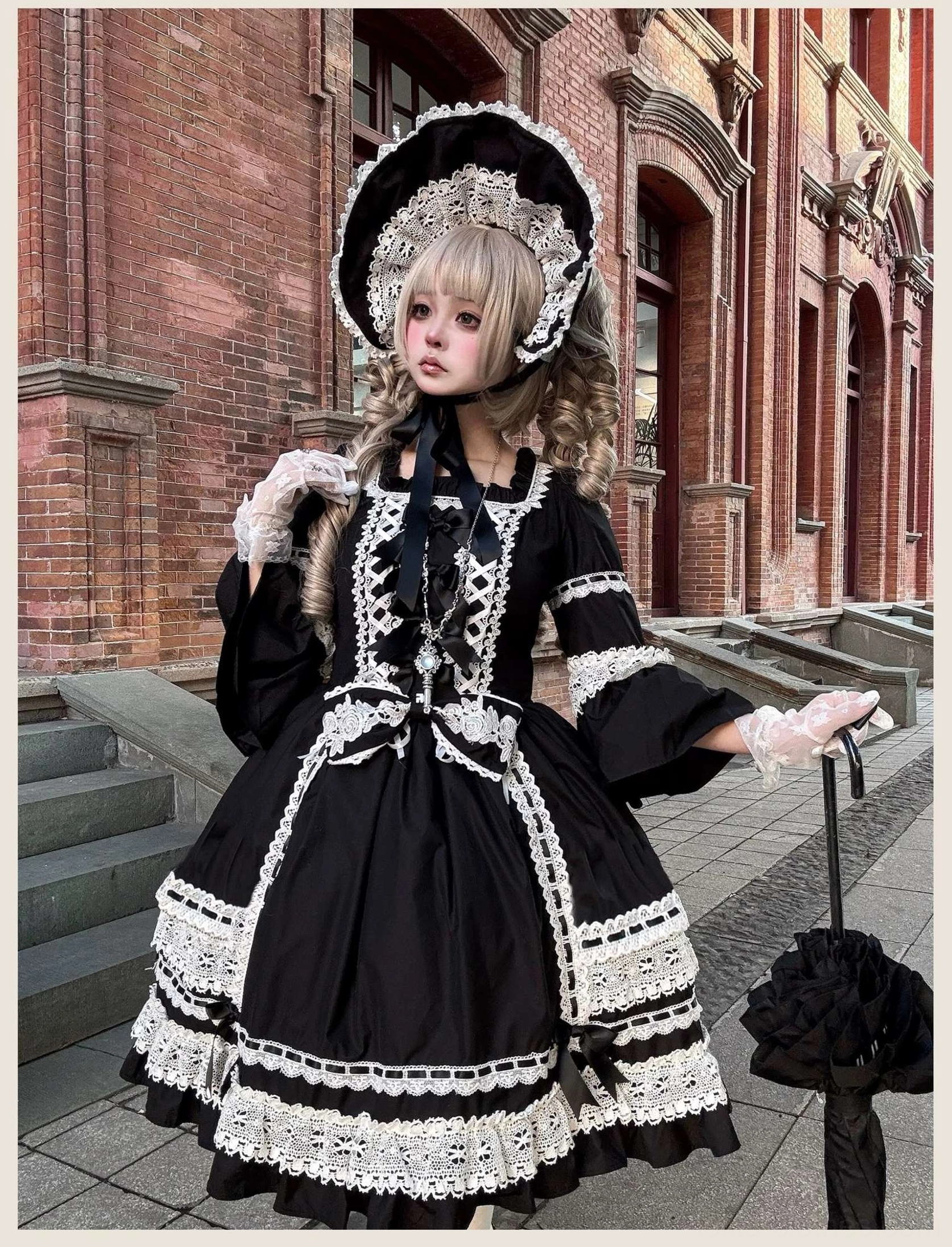 Low angle front view of black vintage old school lolita one-piece dress with full lace trims, black bow decoration, held with black lace umbrella on outdoor brick stairs